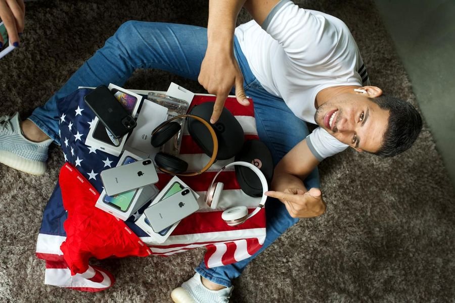 Tyago Sena em 2018, sentado no chão com iPhones e fones importados sobre uma bandeira dos Estados Unidos
