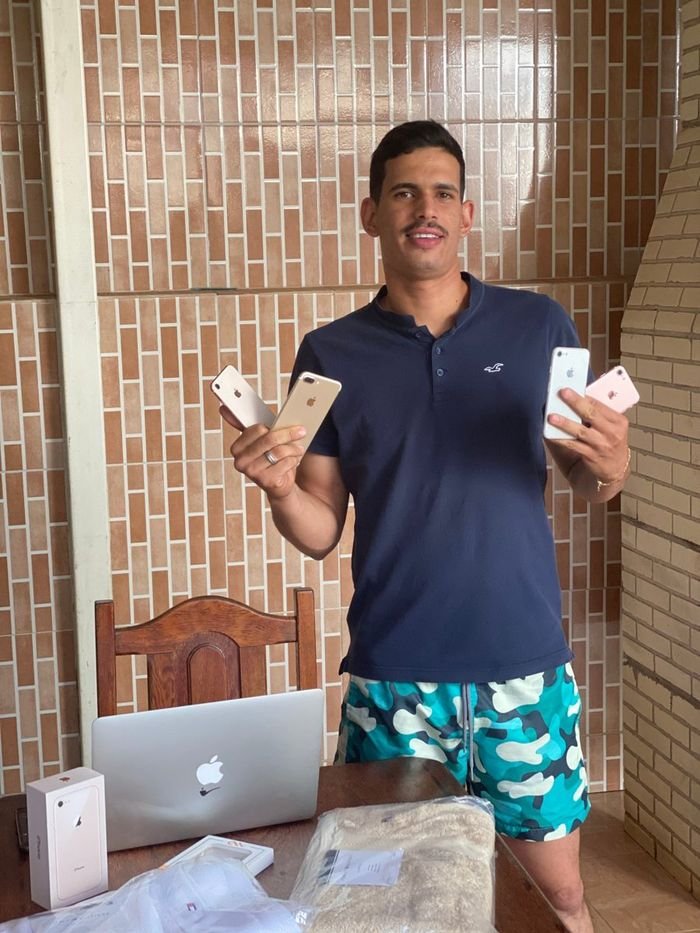 Tyago Sena em casa segurando quatro iPhones, com MacBook e caixas na mesa